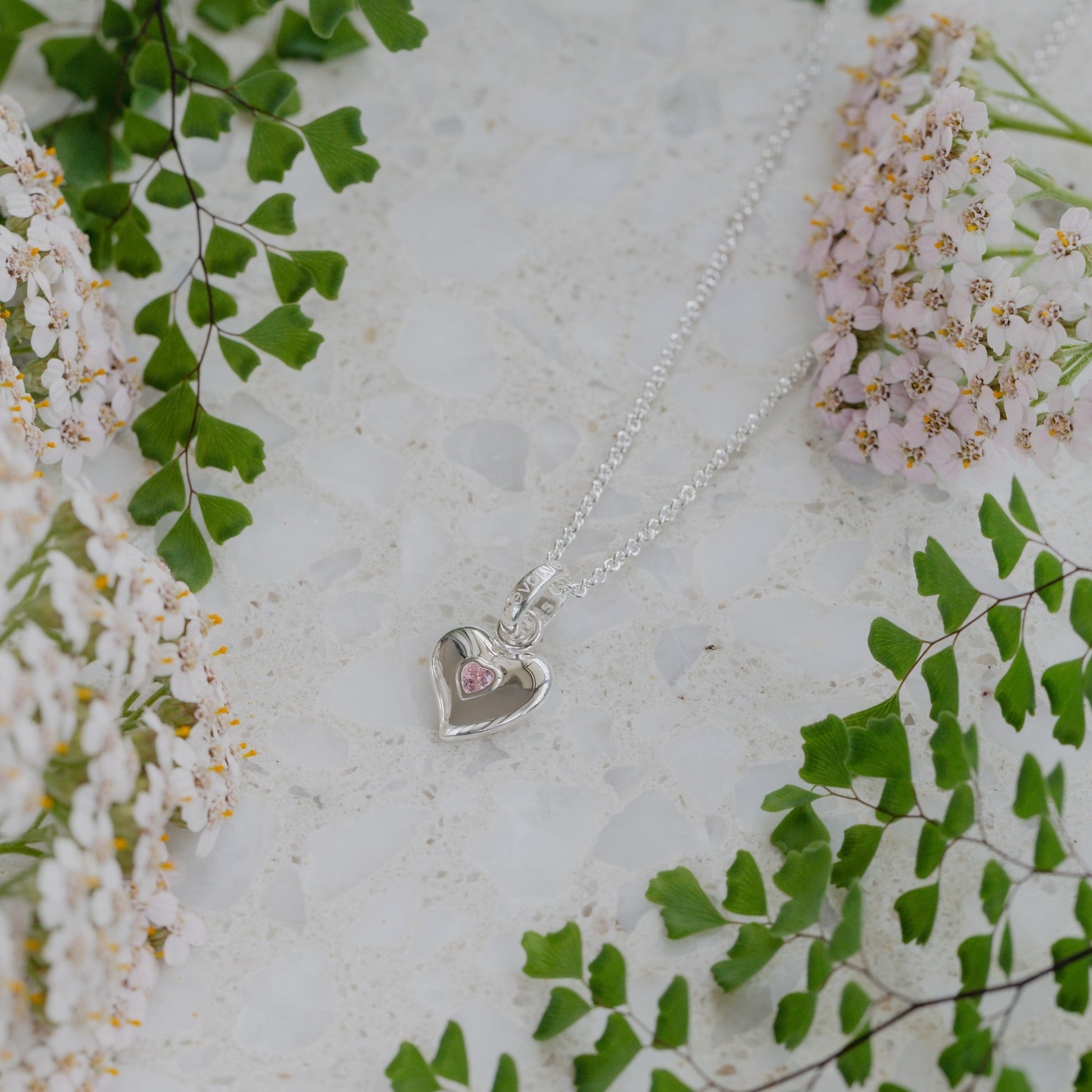 Silver heart-shaped necklace with a pink stone on a light background with green leaves and flowers.