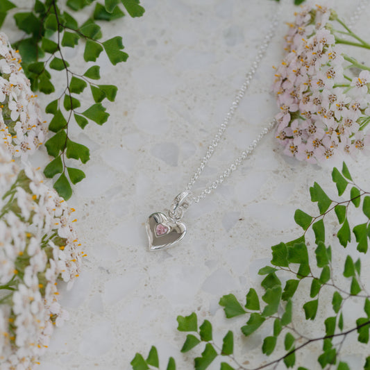 Silver heart-shaped necklace with a pink stone on a light background with green leaves and flowers.