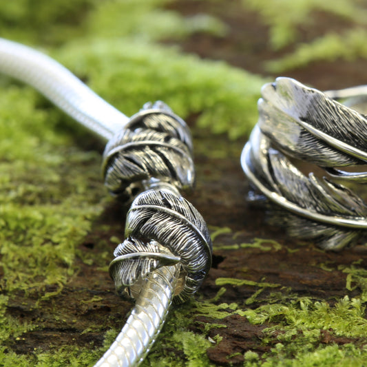 Silver Huia feather charms on a bracelet and ring on a mossy surface